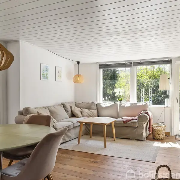 En lys stue med en stor, grå hjørnesofa ved siden af et vindue, der giver udsigt til en have. I nærheden står et rundt spisebord med stole, og der hænger en unik loftlampe. Brændeovn i højre side.