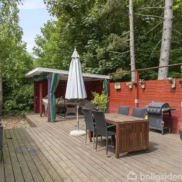 Et rødt hus omgivet af træer og buske ligger i et grønt skovområde. I baggrunden ses en sø og flere huse langs kysten under en blå himmel.
