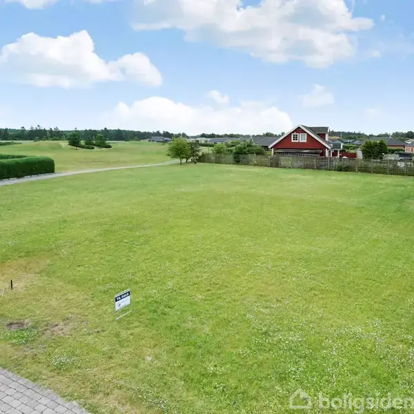 Et stort grønt græsareal strækker sig i forgrunden, med et skilt i venstre side. En rød bygning står i baggrunden omgivet af træer og himlen er let skyet.