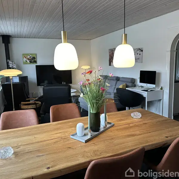 En spisebord i træ dekoreret med blomster står i en stue med sofagruppe og fjernsyn. Moderne lamper hænger over bordet, og der ses en computer i baggrunden.