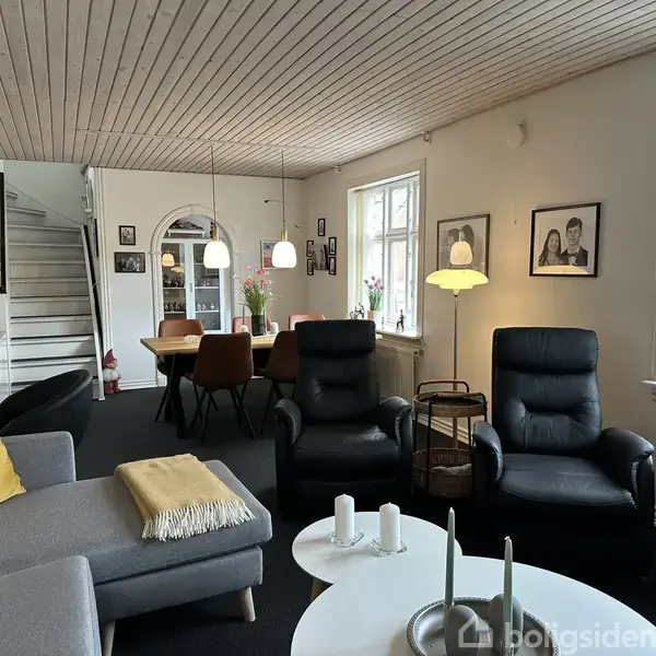 Stue med grå sofa og to sorte lænestole foran et sofabord med stearinlys, i nærheden af en spiseplads med brune stole under hængende lamper og billeder på væggen.