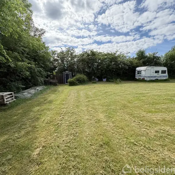 En campingvogn står på en velplejet græsplæne. Omgivet af grønne træer og buske, skaber det en idyllisk og fredfyldt haveatmosfære.