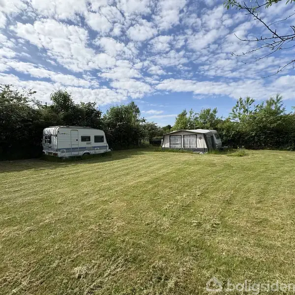 Campingvogn og teltvogn står stille på en velplejet græsplæne omgivet af buske og træer under en blå himmel med lette skyer.