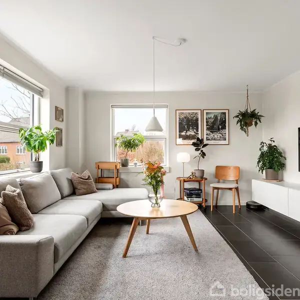 Gråt sofaarrangement med puder står i en lys stue. En rund træbordplade foran sofaen har en vase med blomster. Pyntet med planter og billeder på væggen. En fladskærms-TV er monteret.