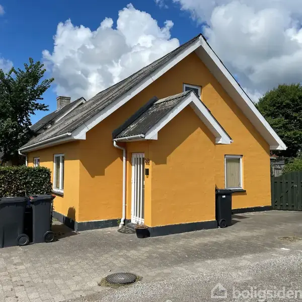 Et gult hus står i en solrig indkørsel med en brostensbelagt sti, omgivet af grønne træer og buske. To skraldespande står tæt ved husets side.