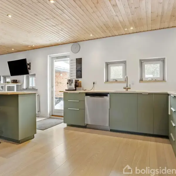 Køkken med grønne skabe og induktionskomfur. Et åbent rum med lyst trægulv og træloft. Flere vinduer lader naturligt lys ind, og en barstol står ved køkkenøen.