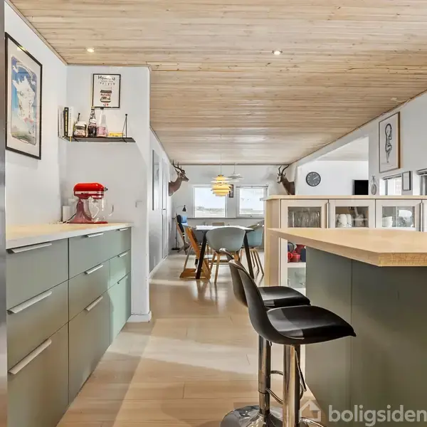 En moderne køkkenø med to sorte barstole står i et lyst køkkenmiljø. Der er vægmonteret TV og hylder med bøger og en rød køkkenmaskine på en skabsoverflade.