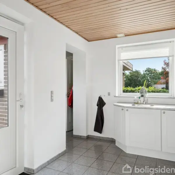 Hvidt køkken med grå fliser og træloft, hvor en vask er indbygget under et vindue. En dør fører udenfor, og der hænger et mørkt håndklæde på væggen.