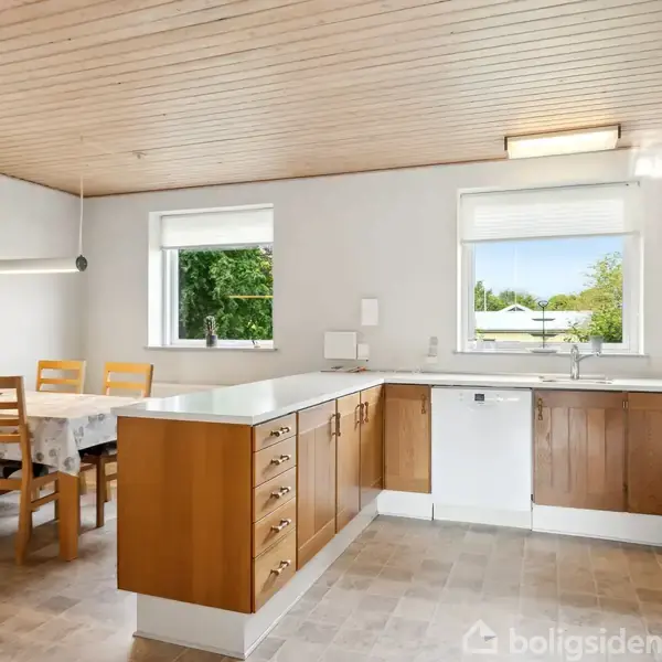 Køkken med træskabe og -skuffer omringer en hvid vask og opvaskemaskine. Spisebord med fire stole og en lampe hænger over det. Vinduer viser udsigt til grønne træer.