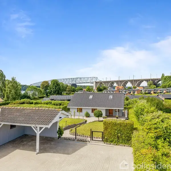 Et hus med en grusbelagt gårdsplads og carport står i forgrunden, mens en stor bro strækker sig over landskabet i baggrunden. Omkring er der frodige grønne buske og træer.
