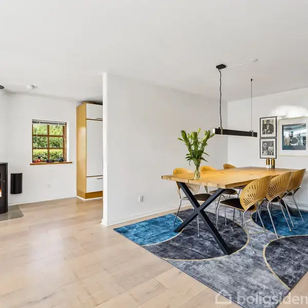 Brændeovn står i hjørnet og brænder roligt i et lyst rum. Spisebord med seks stole ved siden af blåt tæppe. Store vinduer og åben trappe i baggrunden.