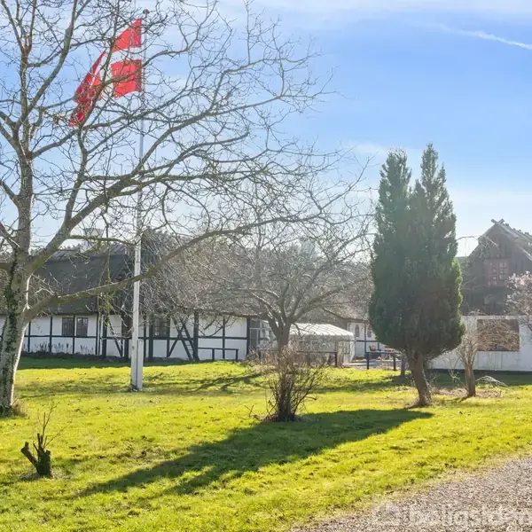 Et bindingsværkshus står stille i solrigt vejr omgivet af græsplæne og spredte træer. En rød dansk flag vajer i vinden fra en flagstang foran huset.
