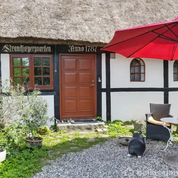 Et bindingsværkshus med stråtag har en rød dør, flankeret af vinduer. En rød parasol skygger over et cafésæt i gruset haveområde. Tekst: "Strandjægergaarden Anno 1787".