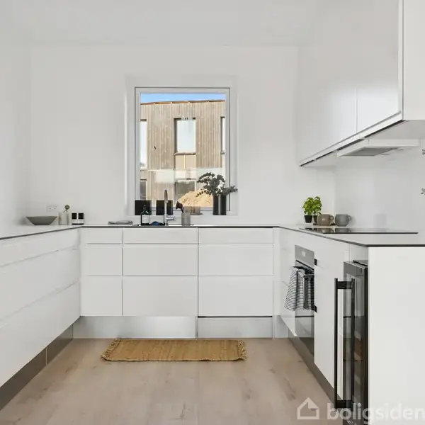 Hvidt køkken med moderne møbler arrangeret langs vægge; ovn indbygget i skab. Et vindue med udsigt udenfor, planter og dekorative genstande. Lyse trægulve kompletterer rummet.