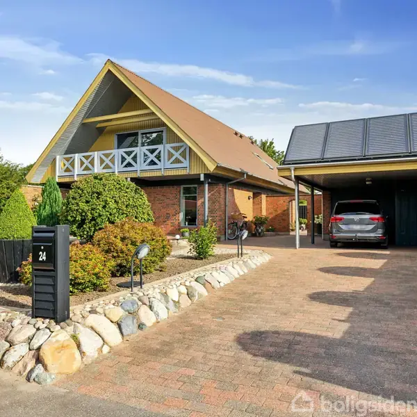 Et moderne hus med A-formet tag står ved siden af en overdækket carport med en bil. Forhaven er pyntet med sten og buske. Husnummeret 24 vises på en sort postkasse.