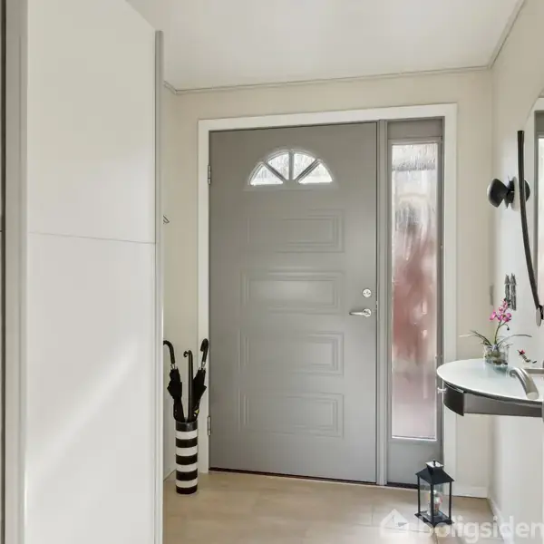 En grå dør står foran en entré med lyse vægge. Til venstre ses en hvid skabsvæg, og til højre en sort-hvid paraplybeholder samt et lille bord med blomster.