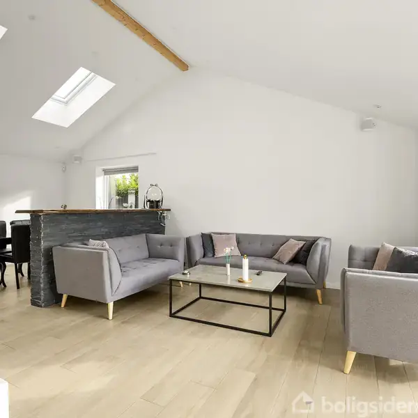 Gråt møbelsæt arrangeret omkring et glas-sofabord; åbent rum med loftsvinduer og træbjælker; spisebord og stole i baggrunden nær dør til have.