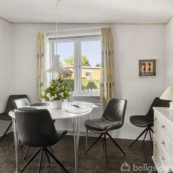 Et rundt bord omgivet af fire sorte stole står i et lyst rum. Bordet har en vase med blomster. Et vindue med gardiner lukker dagslys ind, og kunst hænger på væggene.