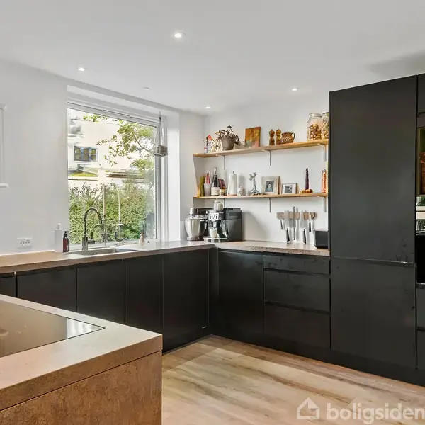 Køkken med sorte skabe og træhylder, hvor en kaffemaskine og køkkenredskaber er placeret. Et stort vindue giver udsigt til haven. Lyst trægulv og loftlys skaber en moderne atmosfære.