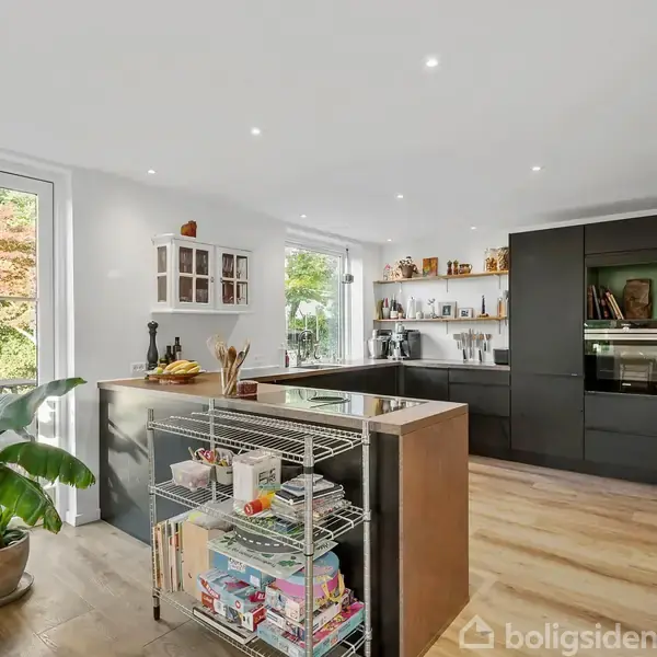 Et moderne køkken med mørke skabe og indbygget ovn. Et metalstativ fyldt med bøger står midt i rummet. Store vinduer slipper lys ind og afslører en frodig have udenfor.