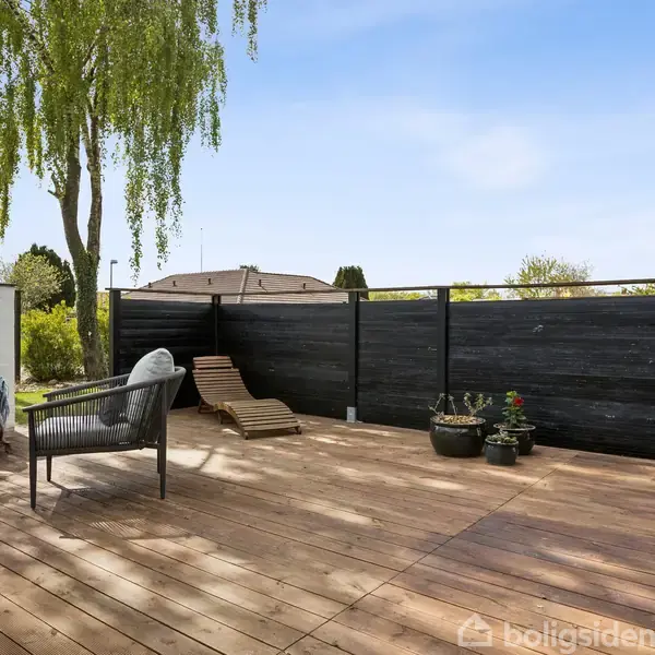 En terrasse med træplanker har havemøbler og potteplanter. Et hegn omkranser området, og der er en stor parasol. I baggrunden ses en frodig have med træer og græsplæne.