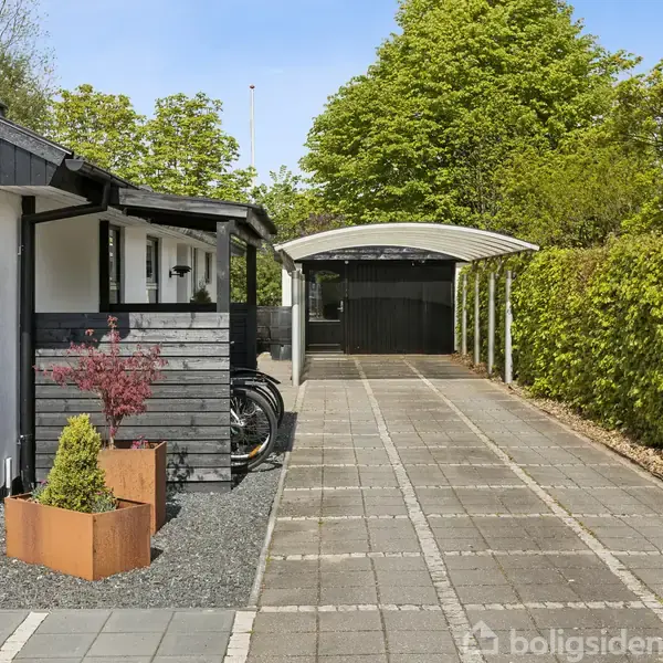 Et hus med nummeret 161 har en indkørsel med fliser og en overdækket carport. Omkring er der grønne træer og buske, og forhaven er pyntet med små buske i plantekasser.