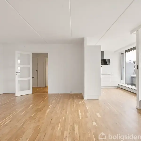 Et lyst og tomt rum har trægulv, en åben dør, og store vinduer mod højre giver udsigt til en balkon. I baggrunden ses et køkken med hvide elementer og emhætte.