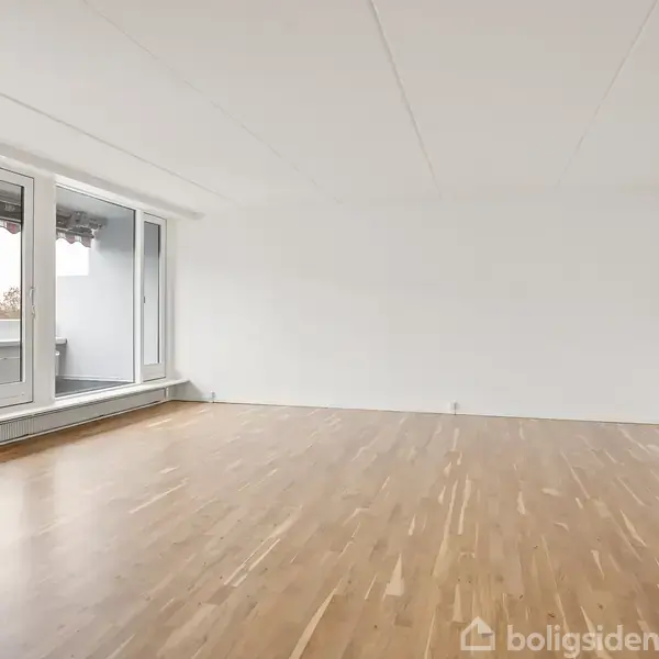 Et rummeligt tomt værelse med lyst trægulv. Store vinduespartier på venstre væg giver udsigt til en udendørs balkon, hvilket skaber en lys og åben atmosfære.