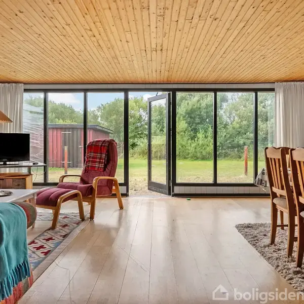 Stue fyldt med lyse træmøbler, en rød- og blåstribet sofa, TV på et bord, og spisestuebord til højre. Store glasdøre åbner til grøn have.