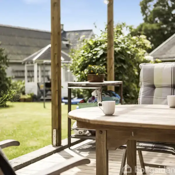 Et rundt havebord med to hvide krus står på en terrasse. Sollys oplyser bordet, mens en frodig have og flere huse ses i baggrunden, omgivet af grønne planter.