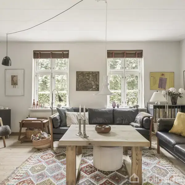 Stue med to sofaer og et sofabord med stearinlys i midten. Væggene er dekoreret med kunst, og store vinduer giver masser af lys. En farverig tæppe dækker trægulvet.