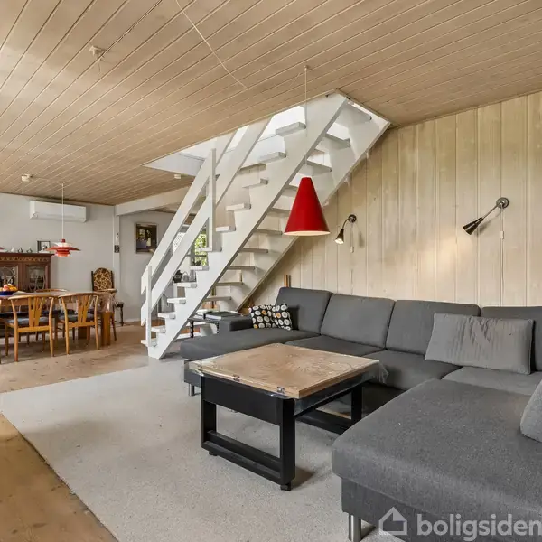 Grå hjørnesofa med sofabord placeret under rød lampe; står i moderne stue med trægulv og trævægge. Trappe fører til øverste etage. Spisebord med stole ses i baggrunden.