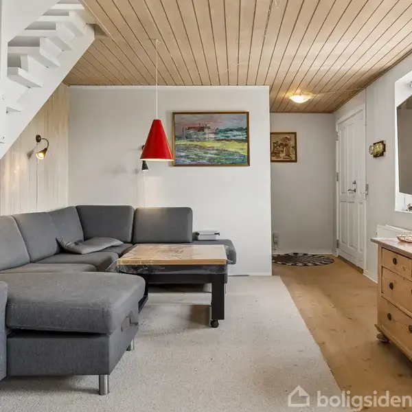 Grå hjørnesofa står ved siden af et sofabord under en rød pendellampe. Trætrapper fører opad. Rummet har lyse trævægge og -loft, og en kommode står ved spejlet.