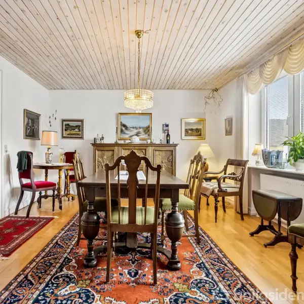 Spisebord med stole står i et elegant rum med trægulv og loft. Et orientalsk tæppe ligger under bordet. Rummet er lyst med store vinduer og klassiske møbler.