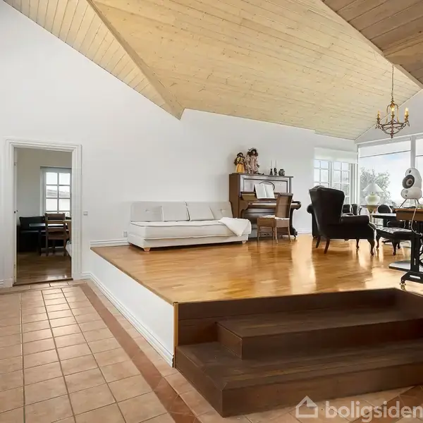 Et rum med trægulvə hævet over fliser, møbleret med sofa, lænestol og klaver. Store vinduer giver masser af lys. Elegant lysekrone hænger fra loftet.