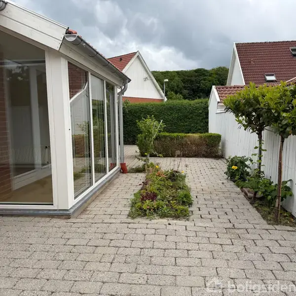 Køkken-alrum med moderne hvidt køkkenbord fyldt med køkkengenstande, tilstødende spiseplads med sort spisebord og stole, store vinduer i baggrunden, der slipper naturligt lys ind.