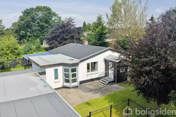 En lav, hvid murstensbygning med fladt tag står stille i en grøn have omkranset af træer. En lille veranda med trin fører til hoveddøren.