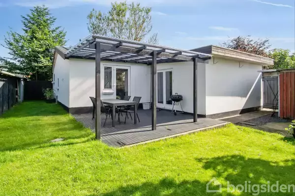 En hvid bungalow med en overdækket terrasse, hvor der står et bord med stole. En grill er på terrassen. Græsplæne og træer omgiver området.