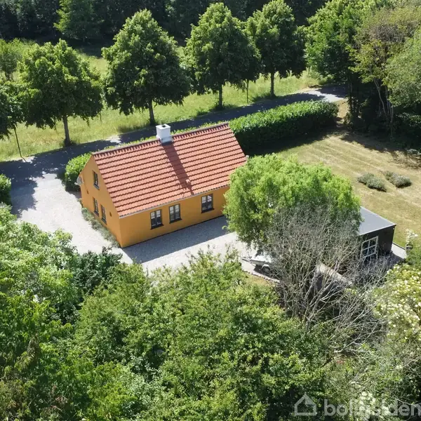 Et gult hus med rødt tag står omgivet af tæt bevoksning og græsplæner. En indkørsel fører op til huset, der ligger i en skovklædt, landlig beliggenhed.