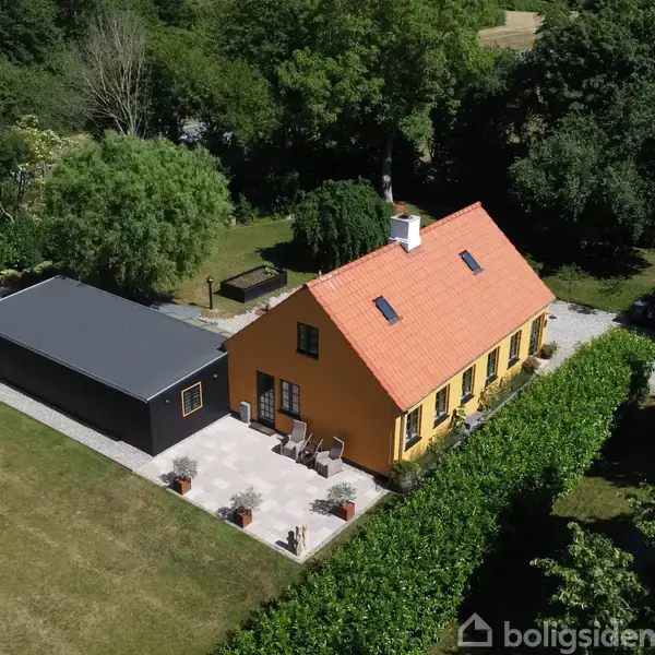 Gult hus med rødt tag står stille ved siden af sort tilbygning. Stor terrasse omkranset af grønne buske og træer omgiver ejendommen i et frodigt landskab.