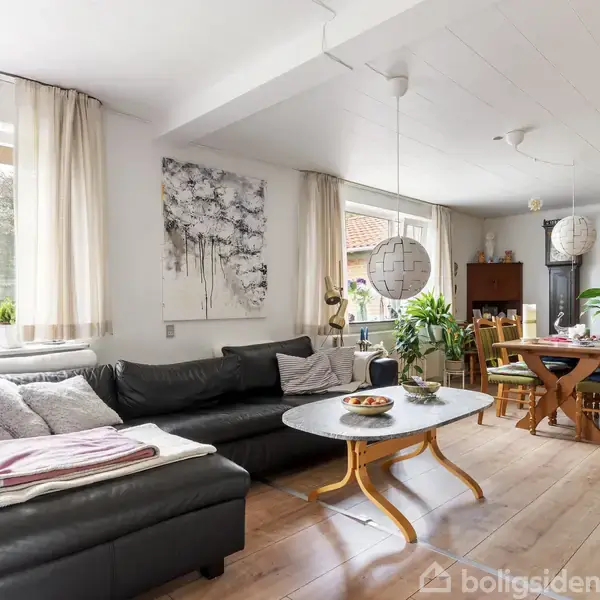 Sort lædersofa med puder og tæpper står i en lys stue. Træbord foran sofaen. spisebord med stole i baggrunden, omgivet af planter og kunst på væggene.
