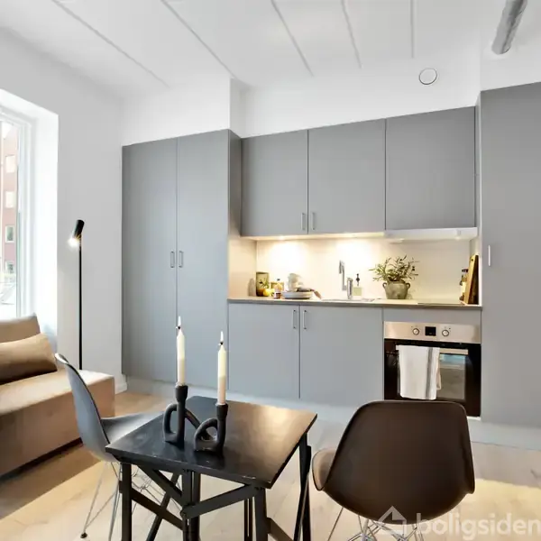 Sofa står i en moderne stue med et lille køkken. Et bord med lys og stole er i forgrunden. Der er lysegrå køkkenskabe og lamper retter lys mod arbejdsområdet.