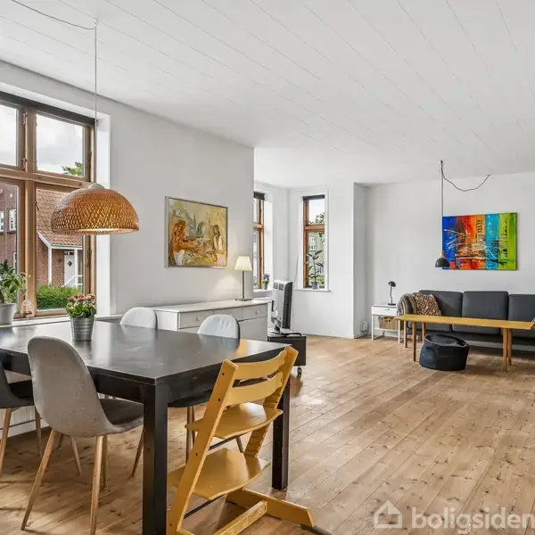 Spisebord med stole, står på trægulv. Ved siden af er der en stue med sofa, sofabord og farverigt maleri på væggen. Store vinduer giver udsigt til have.