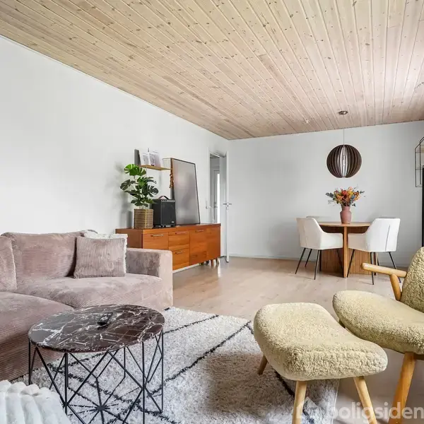 En stue med en beige sofa og et rundt sofabord. Et spisebord med blomster står i baggrunden nær en kommode og hylder med planter. Træloft og lyse vægge.