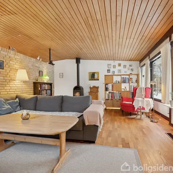 En stue med en stor grå sofa står foran en kaffebord ved siden af en brændovn. Stuen har trægulve, store vinduer, og en væg af mursten.