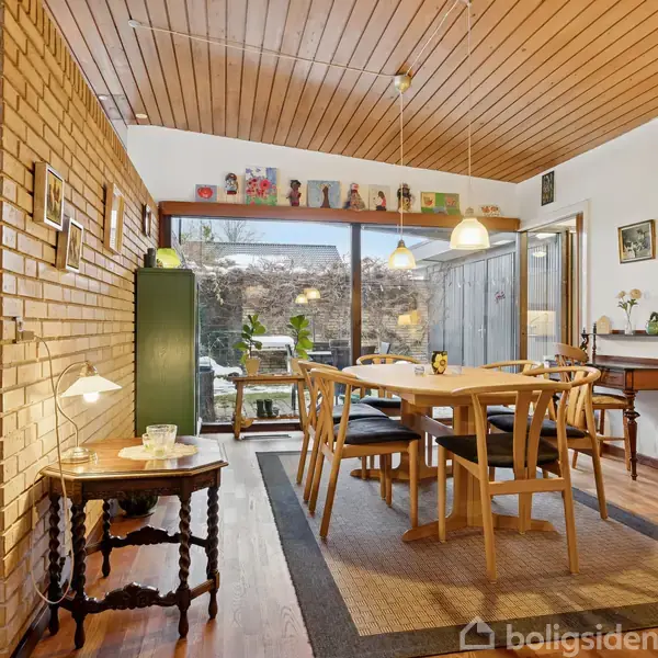 Spisebord står midt i rummet med stole omkring det, oplyst af hængelamper. I baggrunden ses store vinduer, der giver udsigt til en have. Indretningen er klassisk og hyggelig.