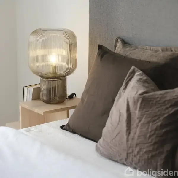 En lampe lyser stilrent på et natbord ved siden af en seng dækket med brune puder. Værelset fremstår moderne og minimalistisk.