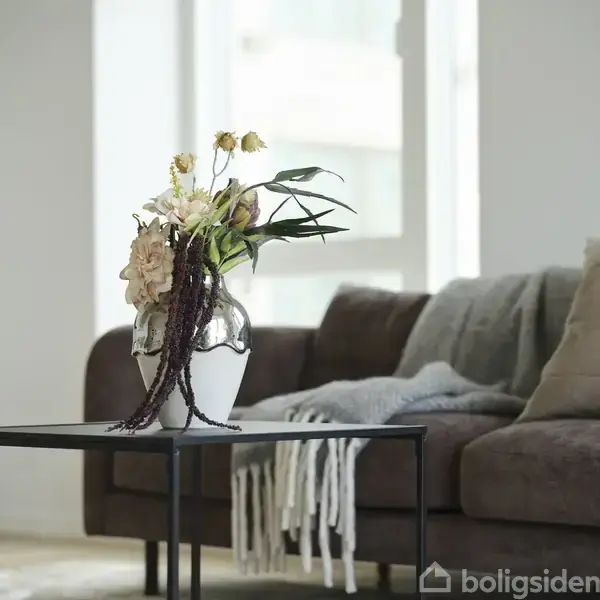 En buket blomster i en vase står på et sort bord. Vase og blomster er placeret foran en brun sofa med puder og et kastetæppe i et lyst rum.