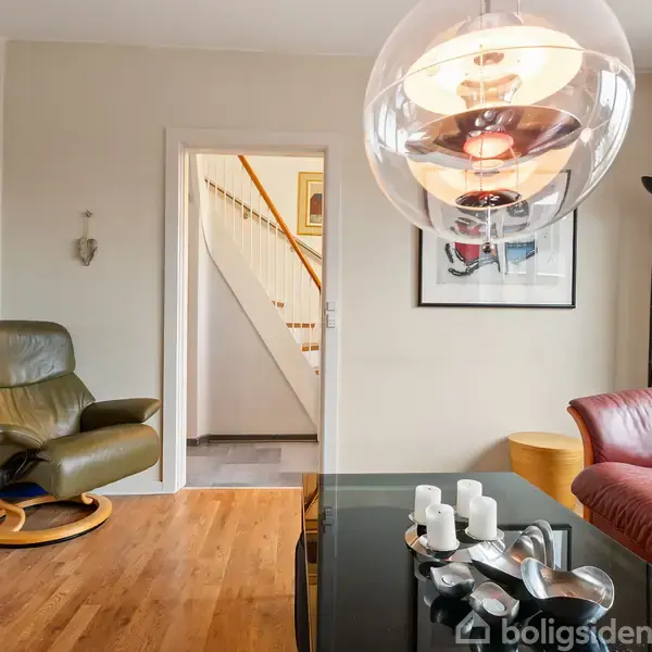 En stue med en grøn lænestol og en rød lædersofa. Et rundt glasbord med dekorative genstande står i midten. En stor lampe hænger over bordet. En trappe ses i baggrunden.