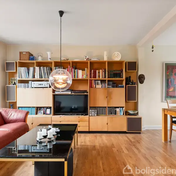 En stue med en rød sofa og sort sofabord, en væg-til-væg bogreol med bøger og fjernsyn samt en spisebordsgruppe med stole. Vinduet slipper naturligt lys ind.